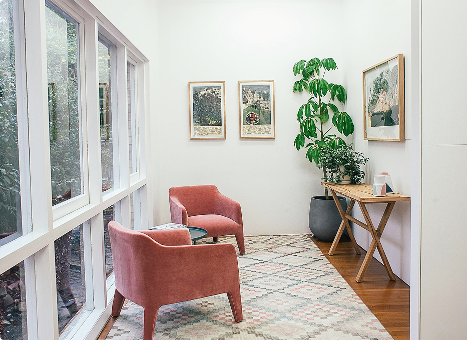 Bright room with two pink chairs, a small round table, framed art on white walls, large leafy plant, and wood console table.